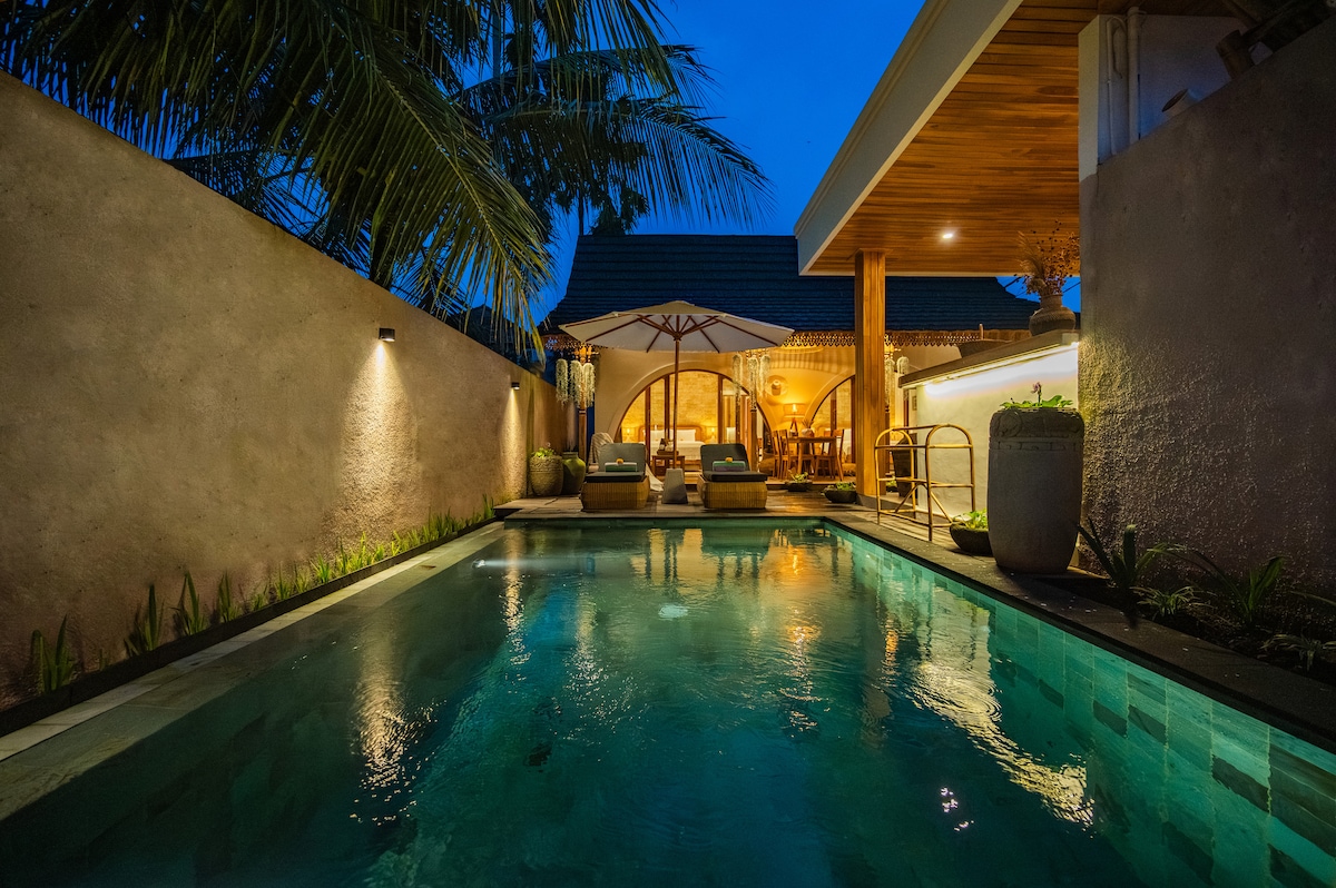 A private pool glows under the evening sky, bordered by lush plants. The tranquil area includes lounge chairs and is framed by the villa's open living space, offering an inviting atmosphere for relaxation and leisure.