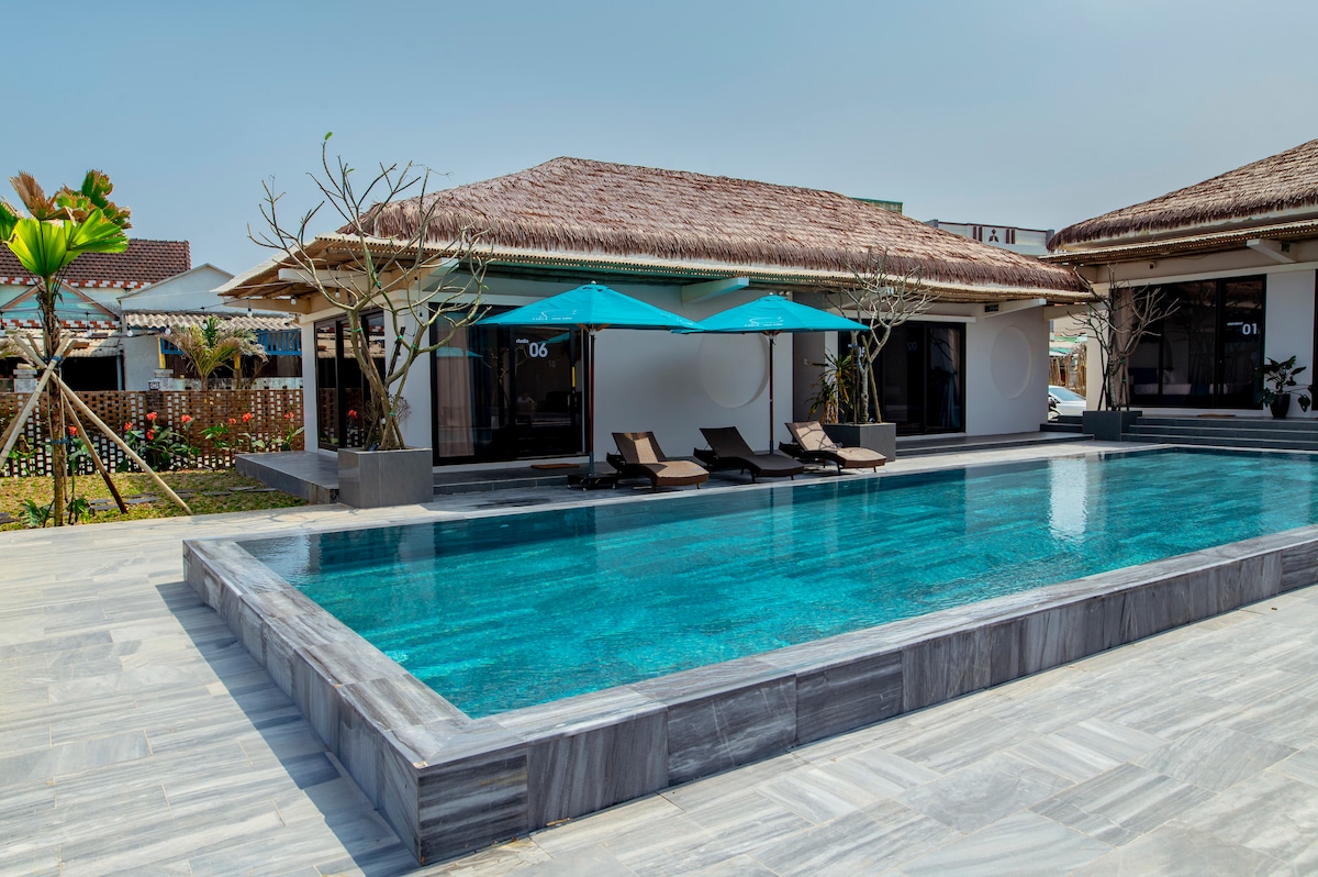 A serene outdoor area features a spacious swimming pool with clear, blue water. Lounge chairs are positioned under large parasols, providing shade. The villa's façade is complemented by a natural thatched roof, while a well-maintained garden is visible in the background.