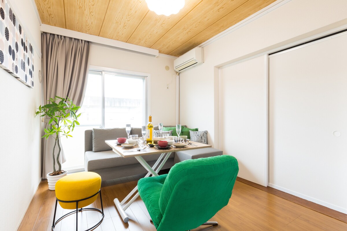 A bright living area features a dining table set for a meal, complemented by colorful chairs. A gray sofa is positioned nearby, while a plant adds a touch of nature. Light streams in through a window, enhancing the overall ambiance of the space.