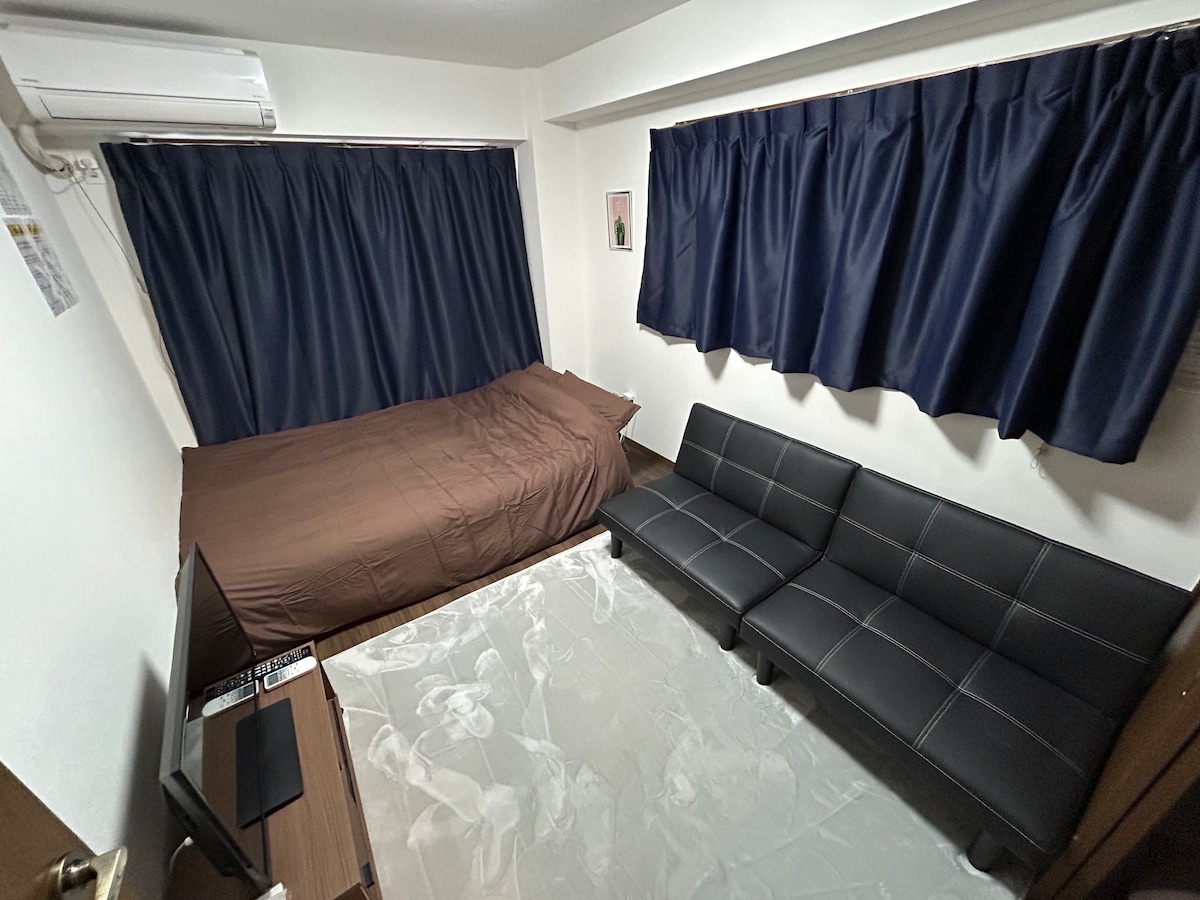 A compact room features a spacious bed covered with a brown duvet. Two sleek black sofas are positioned across from a small television. Dark curtains cover the windows, providing a sense of privacy. A light-colored area rug adds a touch of comfort to the floor.