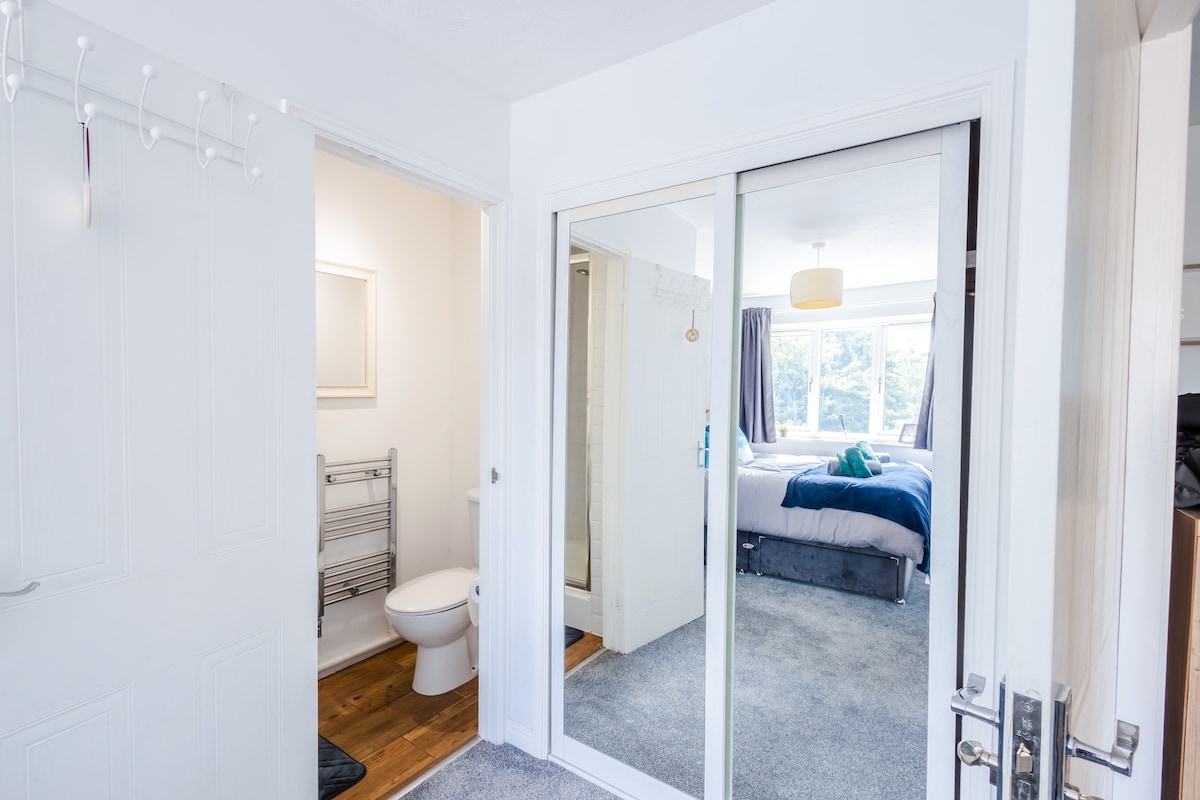 A bright hallway features a mirrored closet door reflecting natural light. To the left, a bathroom is visible with a modern toilet and towel rack. In the background, a bedroom can be seen, showcasing a spacious bed dressed in blue linens and a large window providing outdoor views.