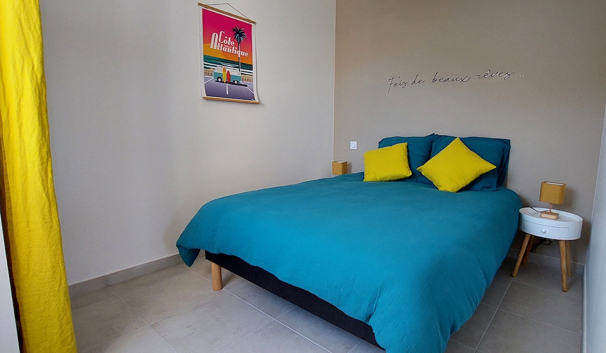 A sleeping area is presented with a spacious bed draped in a teal coverlet. Two bright yellow pillows complement the bedding. Light tones adorn the walls, and a decorative wall hanging adds a touch of character. Natural light enters from a partially opened curtain.