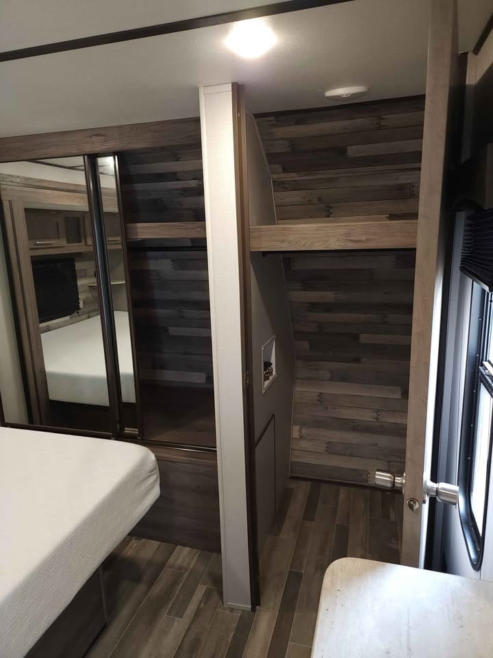 A compact interior showcases a sectioned layout with a bed positioned adjacent to a storage area. The walls feature a subtle wood grain, and natural light enters through a window. The flooring consists of sleek, modern tiles adding to the contemporary feel.