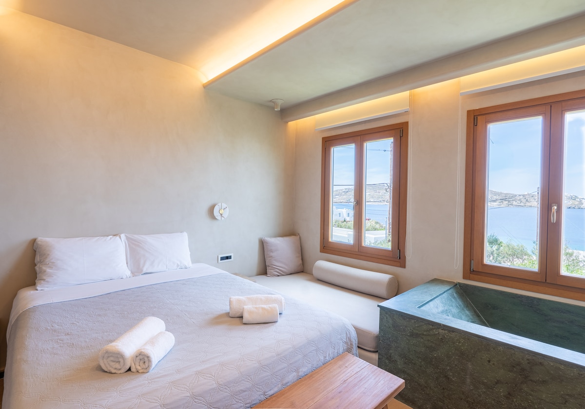 A spacious bedroom features a large bed adorned with fresh linens and neatly arranged towels. A daybed is positioned by the window, providing natural light and views of the nearby water. The soft color palette and simple furnishings create a calming atmosphere.