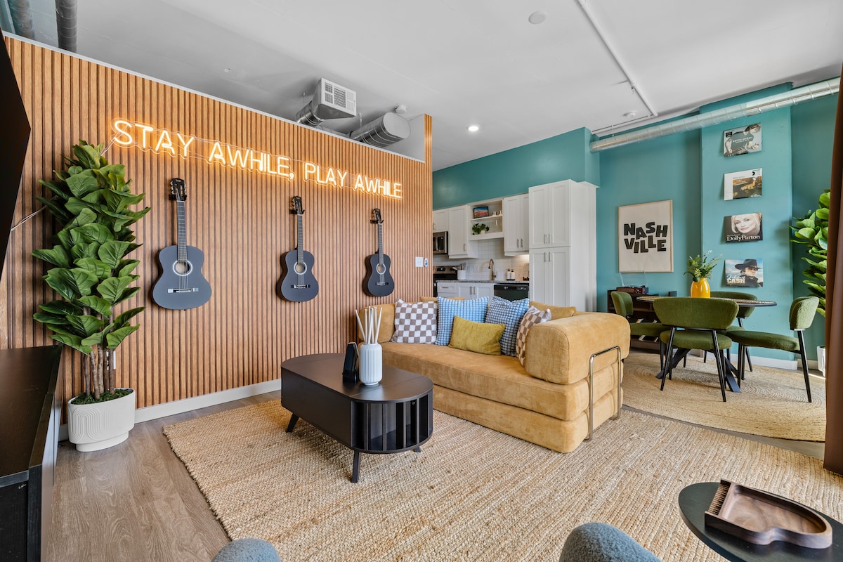 A stylish living area features a modern couch alongside a coffee table. A vibrant green wall offers a backdrop for decorative guitars. The space is illuminated by neon signage, while a dining area is visible in the background, creating a welcoming atmosphere for relaxation or social gatherings.