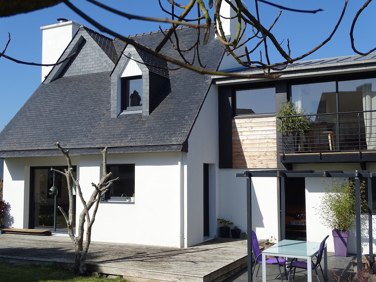 The exterior of the house presents a modern design, featuring a combination of sloped and flat roofs. Large glass windows and a spacious wooden deck are visible, accompanied by a table and chairs. A tree is positioned in the foreground, enhancing the outdoor space.