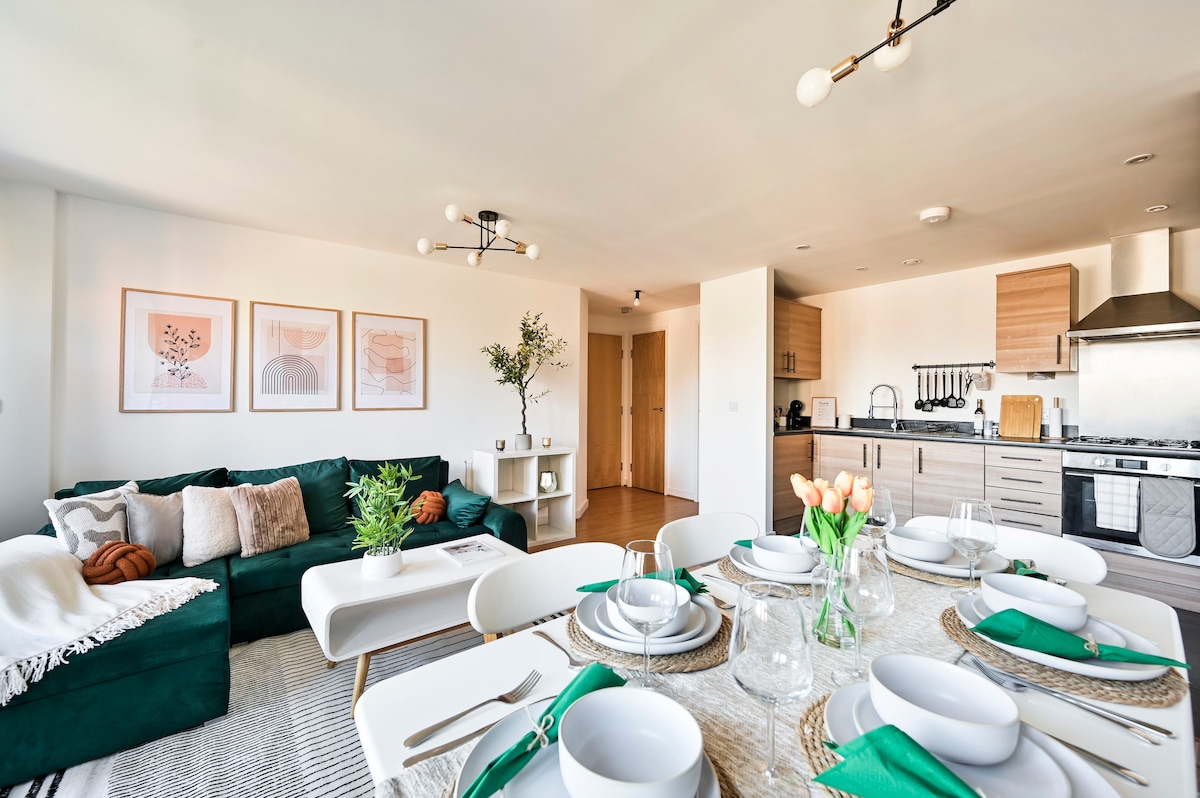 A modern open-plan living area features a large green sofa adorned with textured cushions. A dining table is set with white plates, glassware, and green accents. Art prints hang on the walls, while a kitchen area with wood cabinetry is visible in the background.