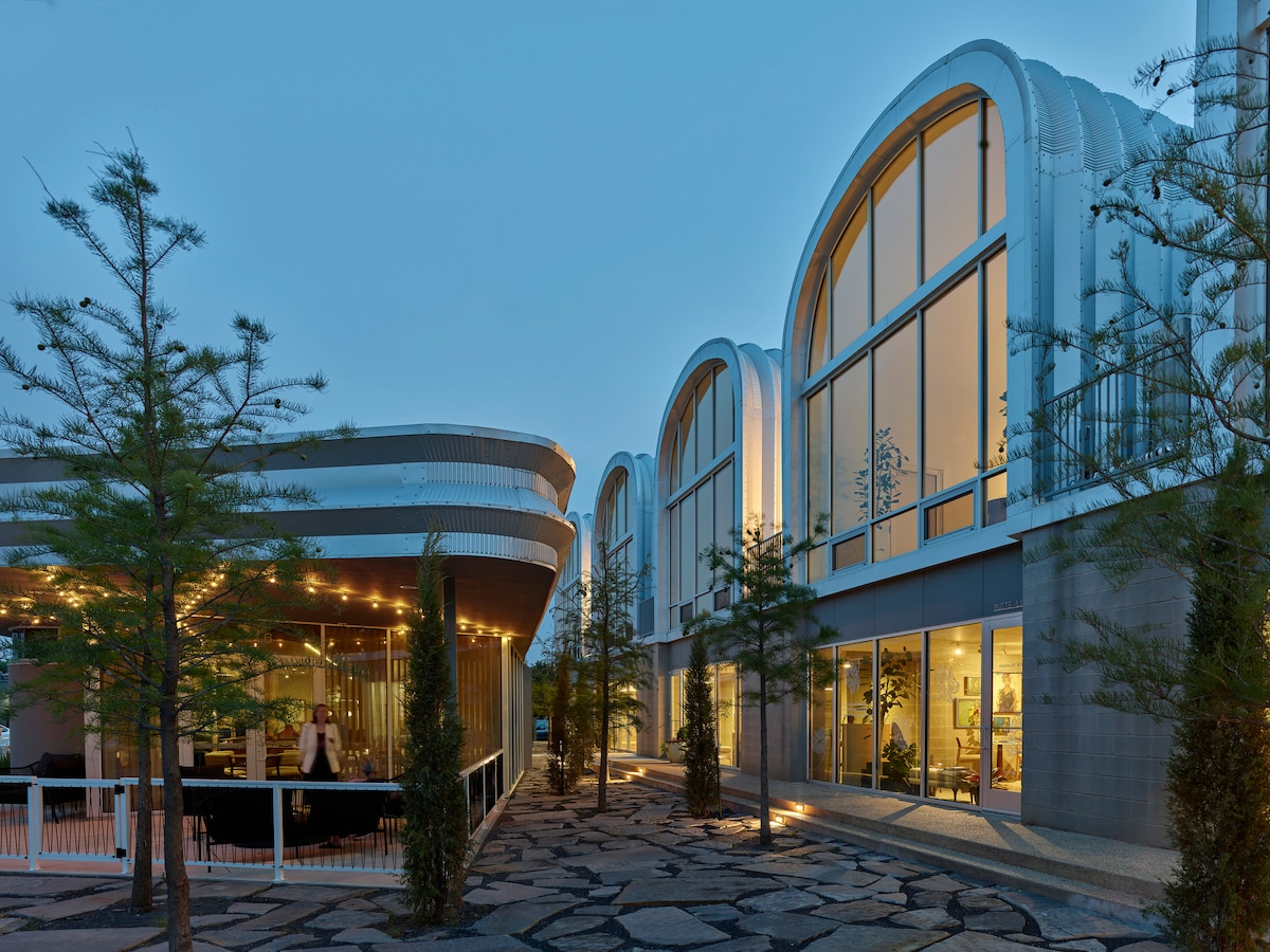The exterior showcases a contemporary architectural design with curving roofs and expansive glass windows. A cozy patio area is highlighted in the foreground, surrounded by neatly arranged shrubs and stones, creating a serene entryway to the vibrant surroundings.