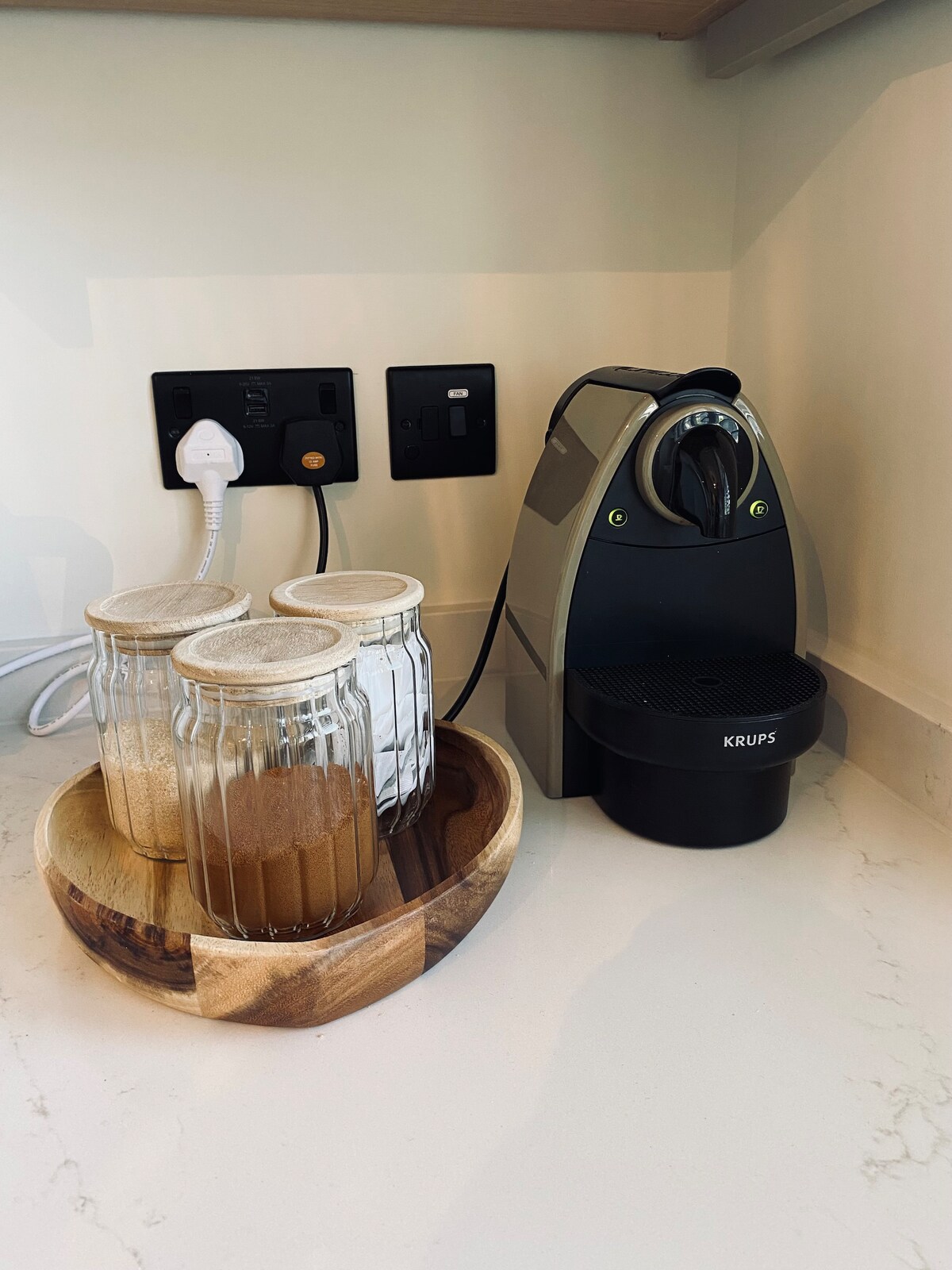 A coffee machine is positioned next to a set of three glass jars filled with various ingredients, all resting in a wooden bowl. The countertop features a clean, modern aesthetic with visible electrical outlets.