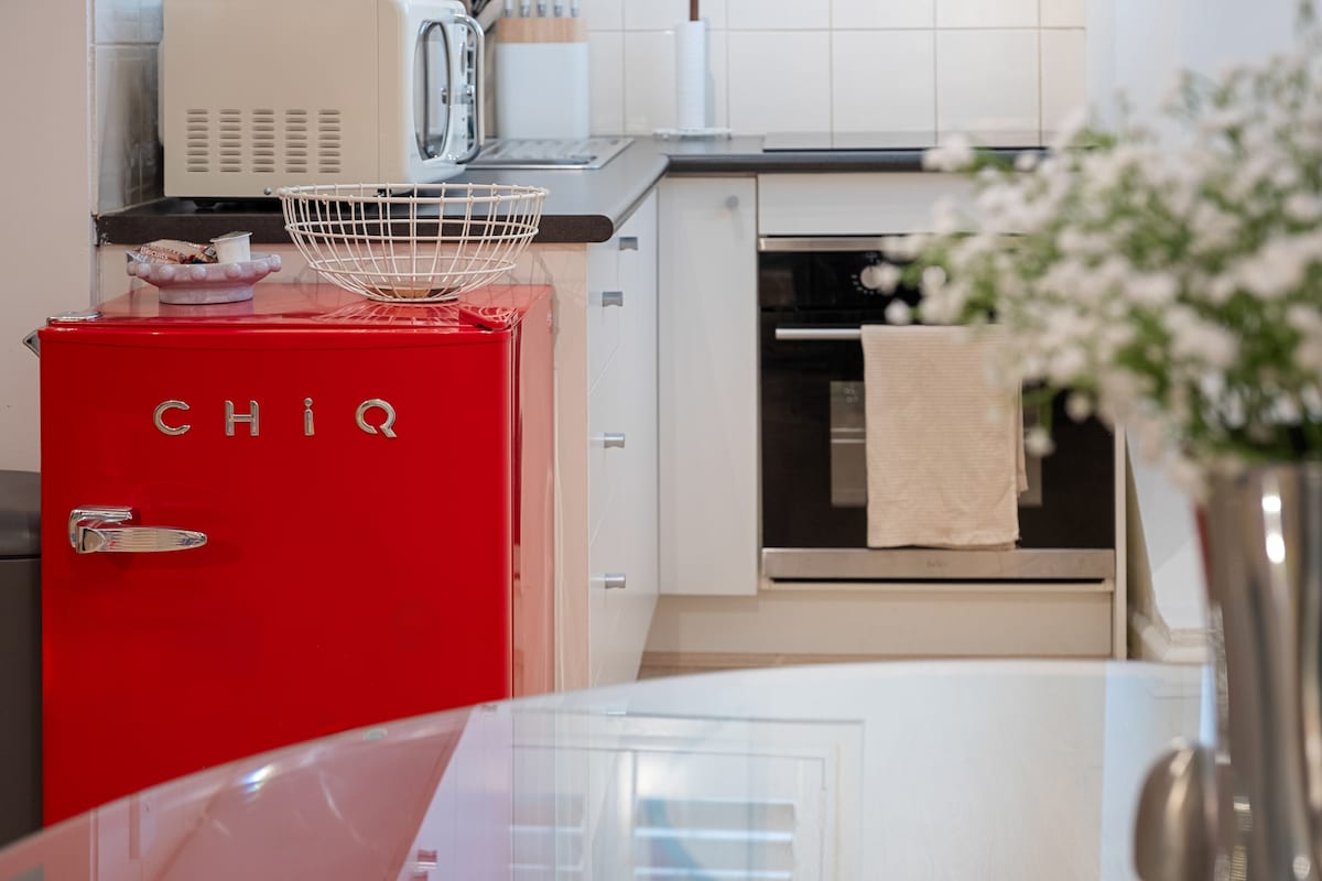 A modern kitchen features a bright red vintage-style refrigerator and sleek white cabinetry. A small countertop microwave is positioned above the stove. A minimalist dining table is visible in the foreground, complemented by a bouquet of white flowers in a metallic vase.