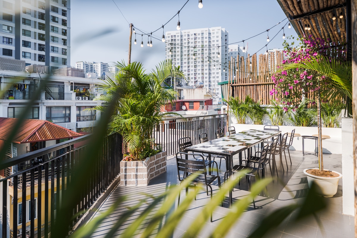 A spacious rooftop area features a large table surrounded by multiple chairs, ideal for gatherings. Green plants and colorful flowers add a touch of nature, while string lights overhead enhance the ambiance. Urban views showcase the surrounding buildings in the background.