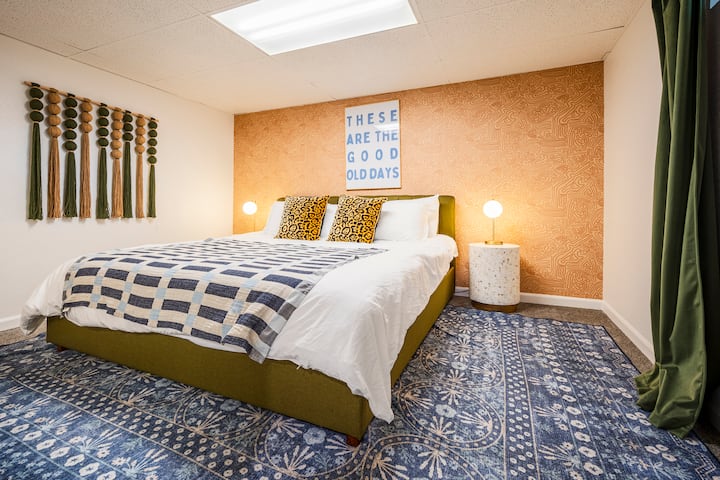 غرفة نوم واسعة مع سرير بحجم كينج، وجدار مميز عصري، ووسائد بنقشة جلد الفهد، ولحاف مريح، وإضاءة دافئة. تتميز بستائر خضراء وفن جداري أنيق وسجادة منقوشة - مثالية لإقامة مريحة ومريحة.