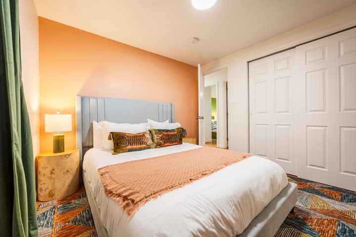 غرفة نوم دافئة وجذابة مع سرير بحجم متوسط، وجدار بلون الخوخ الناعم، ولوح أمامي أزرق فاتح، ووسائد أنيقة، وبطانية مريحة. تتميز بمصباح بجانب السرير ومساحة خزانة وسجادة منقوشة ملونة لإقامة مريحة