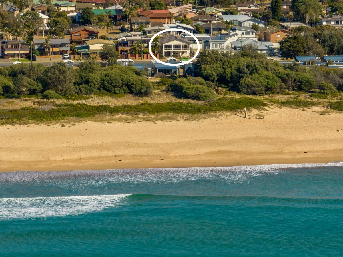 An aerial view captures the coastal property, positioned amongst leafy greenery along the shoreline. The surrounding beach and ocean are visible, along with nearby homes, creating a sense of proximity to both the beach and community amenities.