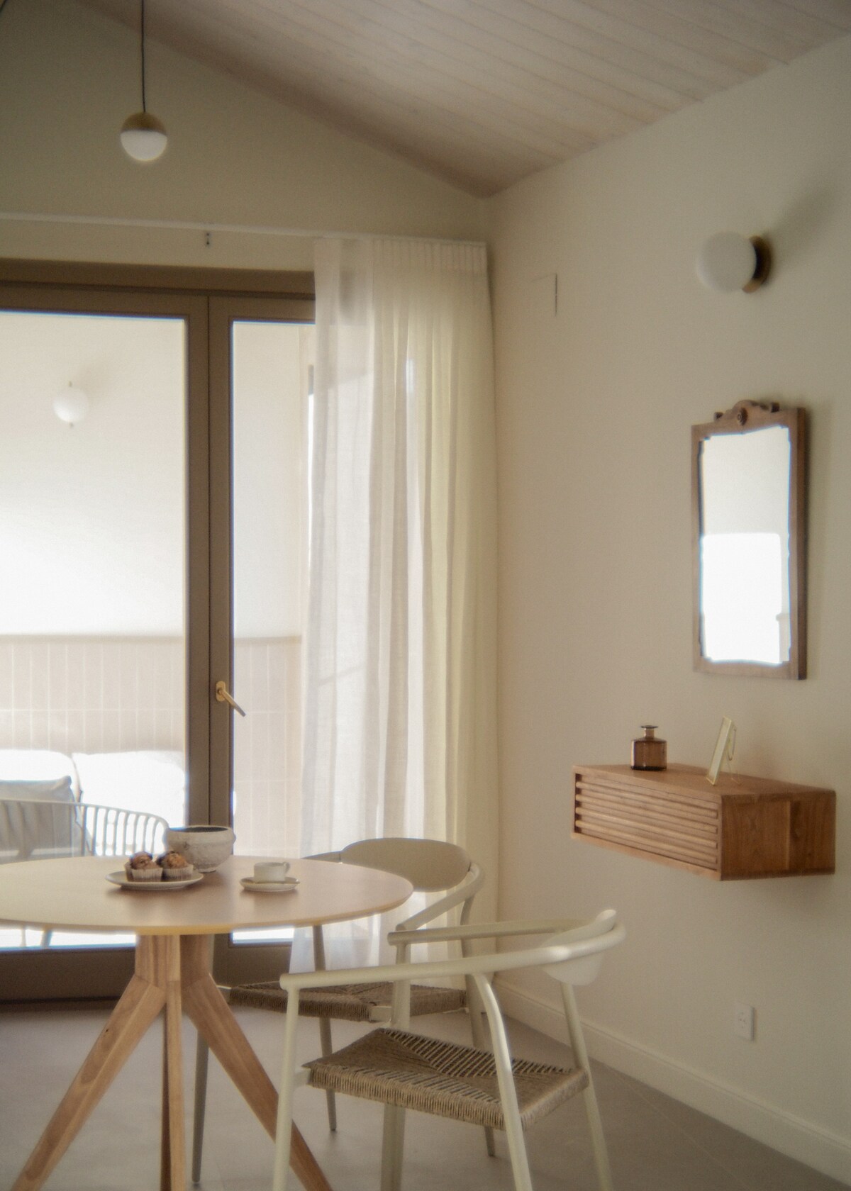 A simple dining area is highlighted by a round wooden table paired with two stylish chairs. Natural light filters through sheer curtains, illuminating the space. A mirror is mounted on the wall above a small wooden shelf, adding functionality while maintaining a minimal aesthetic.
