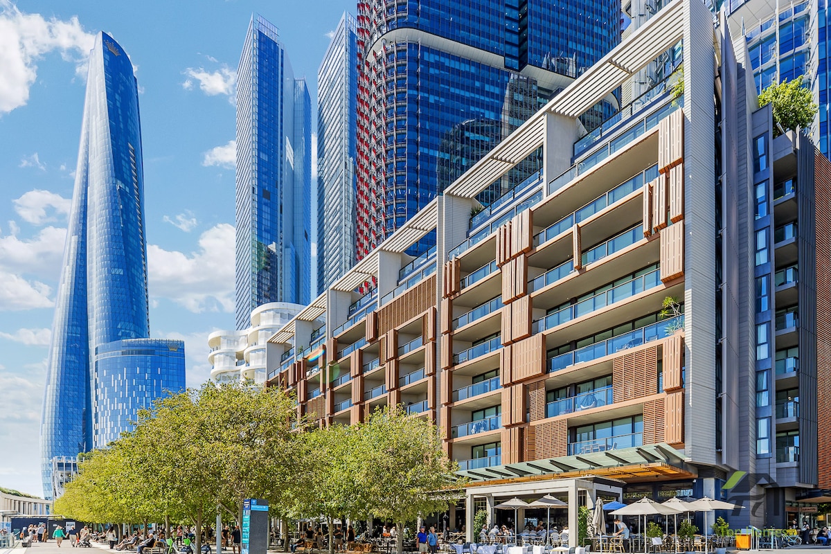A modern urban landscape is displayed, featuring a combination of sleek high-rise buildings and contemporary multi-story accommodations. Lush trees line the foreground, adding greenery to the bustling scene, while outdoor seating areas hint at dining options beneath a bright blue sky.