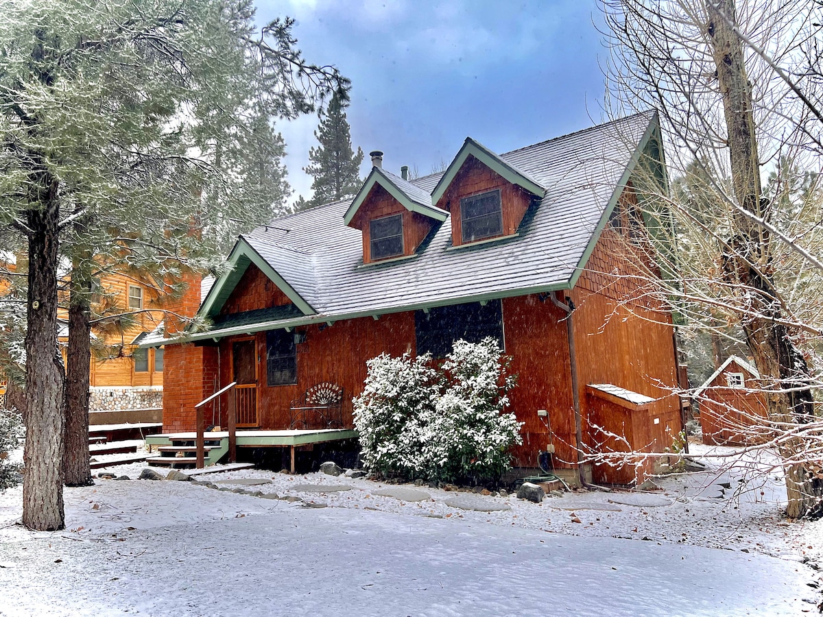 An orange wooden cabin is showcased under a light snowfall, surrounded by a snowy landscape. The steeply pitched roof features dormer windows, and a small porch is seen at the front, bordered by white snow-covered shrubs.