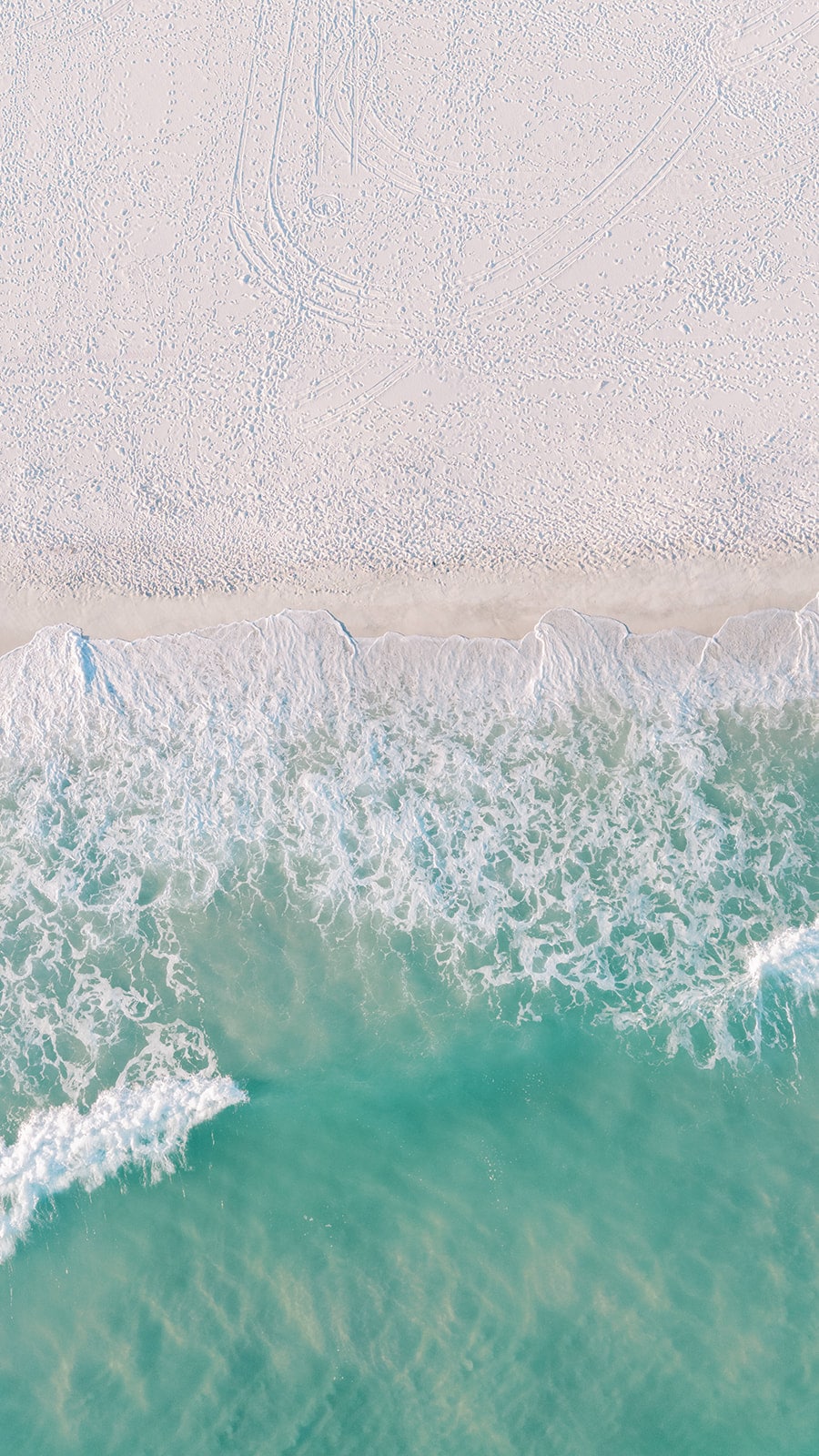 An aerial view captures the shoreline where soft, white sand meets clear, turquoise waters. Gentle waves lap at the beach, creating a serene contrast with the vibrant hues of the ocean, suggesting a tranquil setting perfect for relaxation and leisure.