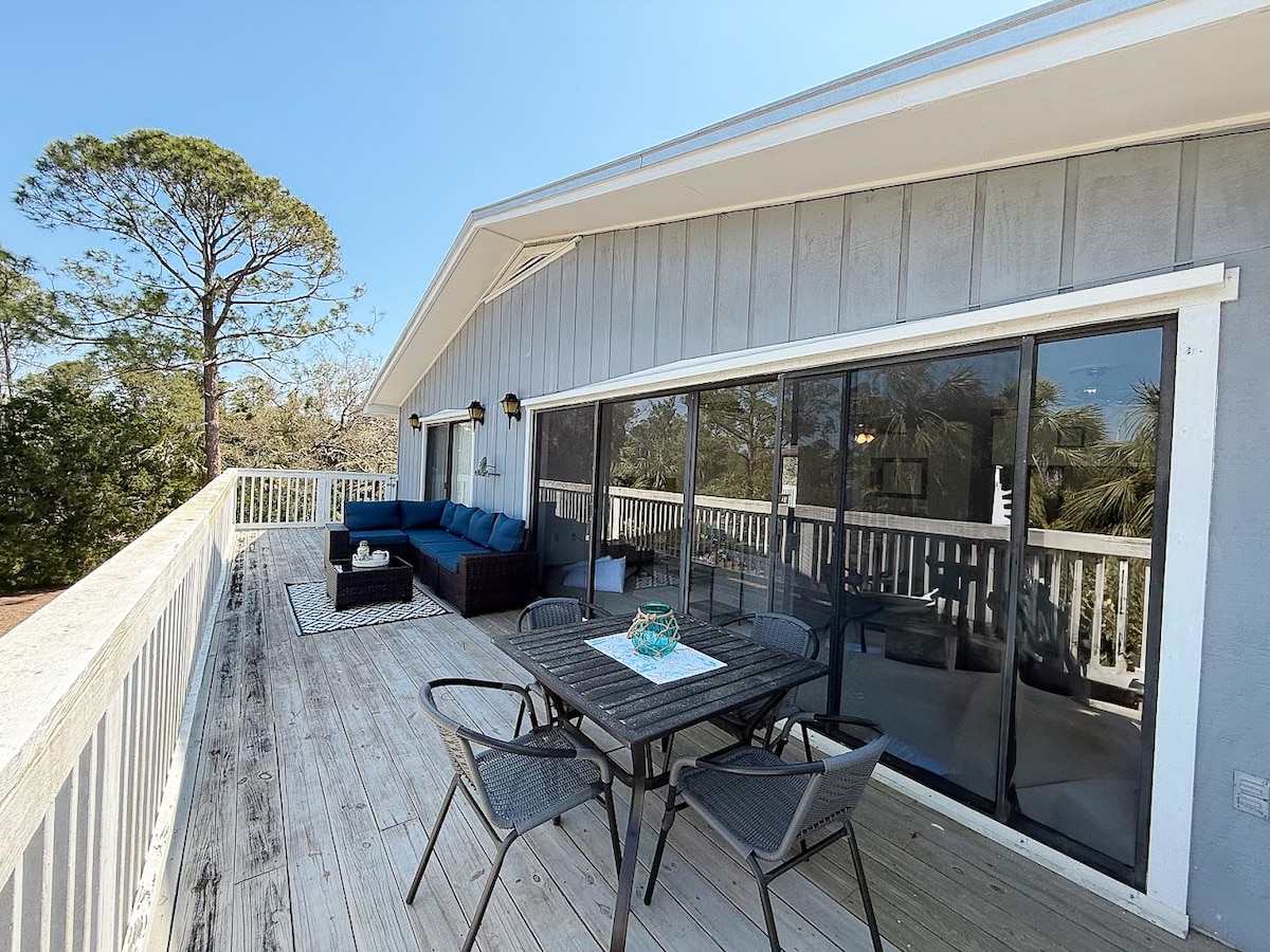 An outdoor balcony features a cozy seating area with a comfortable blue sofa and a dining table for six. Large sliding glass doors offer views of the surrounding greenery, while natural light enhances the inviting atmosphere. The decking provides ample space for relaxation and outdoor dining.