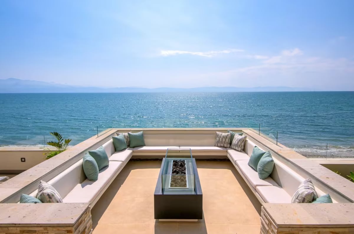 A spacious outdoor lounge area features wraparound seating adorned with light blue cushions, surrounding a glass-topped firepit. The setting overlooks the ocean, showcasing expansive water views under a clear blue sky, with gentle waves lapping at the shoreline.