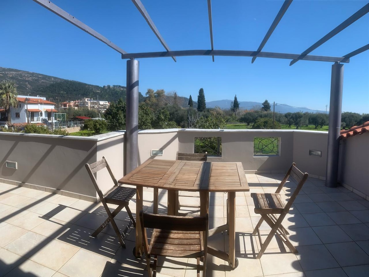 An outdoor balcony space showcases a wooden table surrounded by four folding chairs. A metal frame supports a shaded area above. The view includes distant mountains and greenery, with clear blue skies creating a bright ambiance.