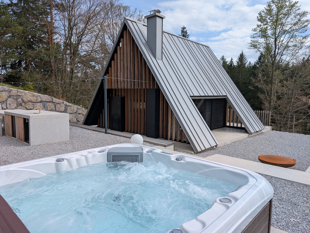 A modern cabin features an A-frame design with a metal roof, surrounded by natural greenery. In the foreground, a hot tub with bubbling water invites relaxation, while a stone terrace extends around the structure, enhancing the outdoor space.