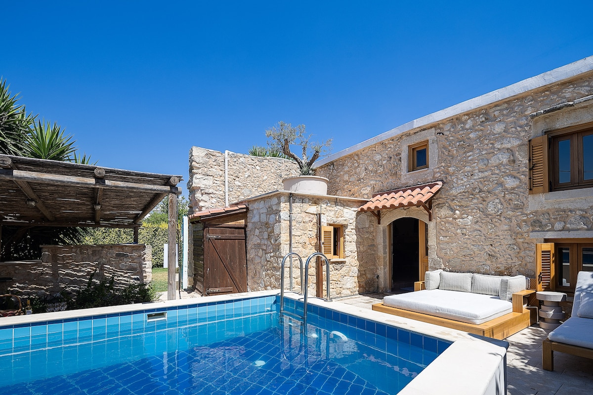 A stone cottage is highlighted amidst a clear blue sky, featuring a private swimming pool with steps for easy access. A wooden lounge area is positioned by the pool, accompanied by sunbeds and a shaded pergola. The rustic architecture adds charm to the outdoor space.