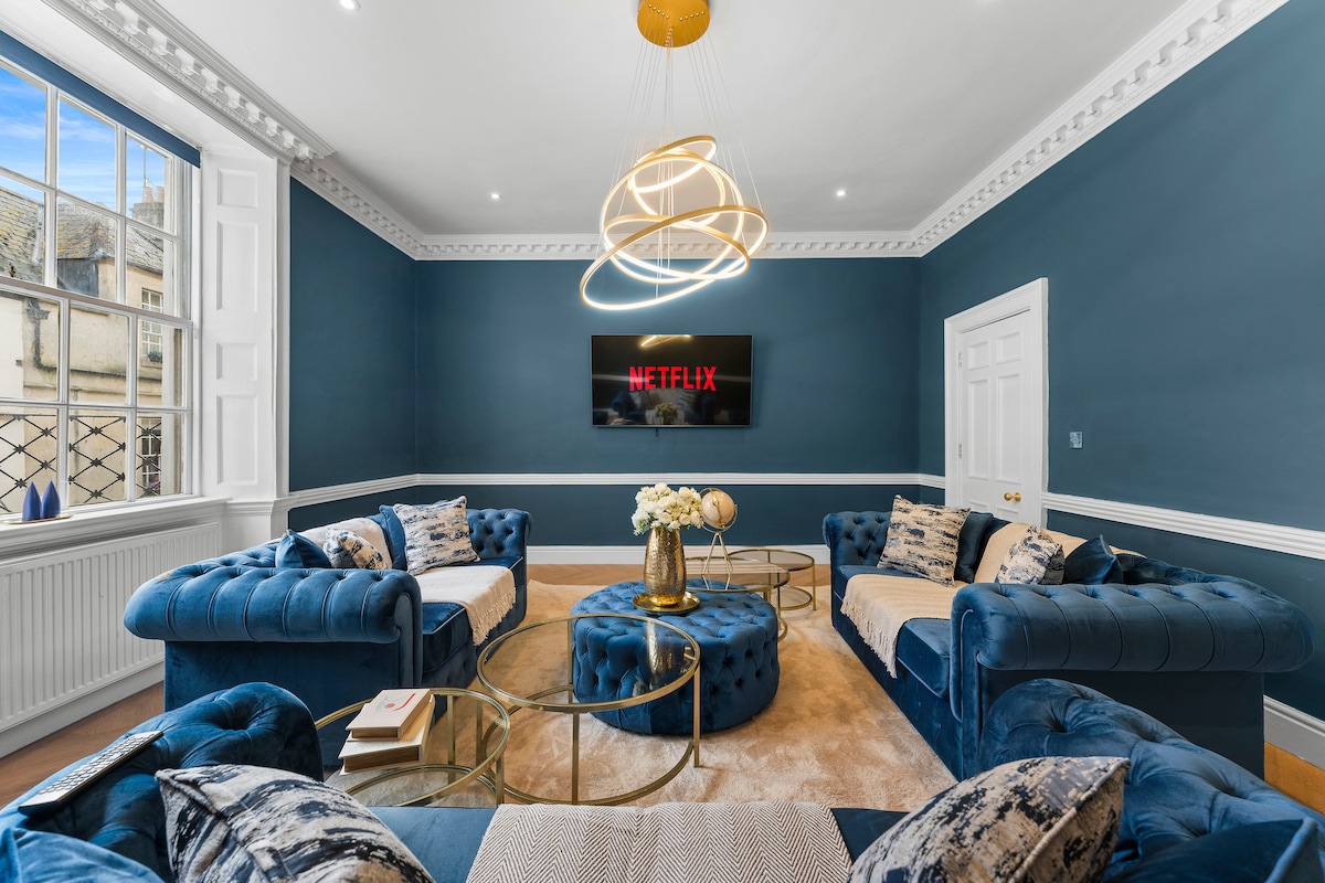 The spacious living room features deep blue walls and elegant ceiling details. Three chesterfield sofa beds are arranged around a central glass table, complemented by a soft area rug. A modern light fixture hangs above, and a wall-mounted screen displays the Netflix logo.