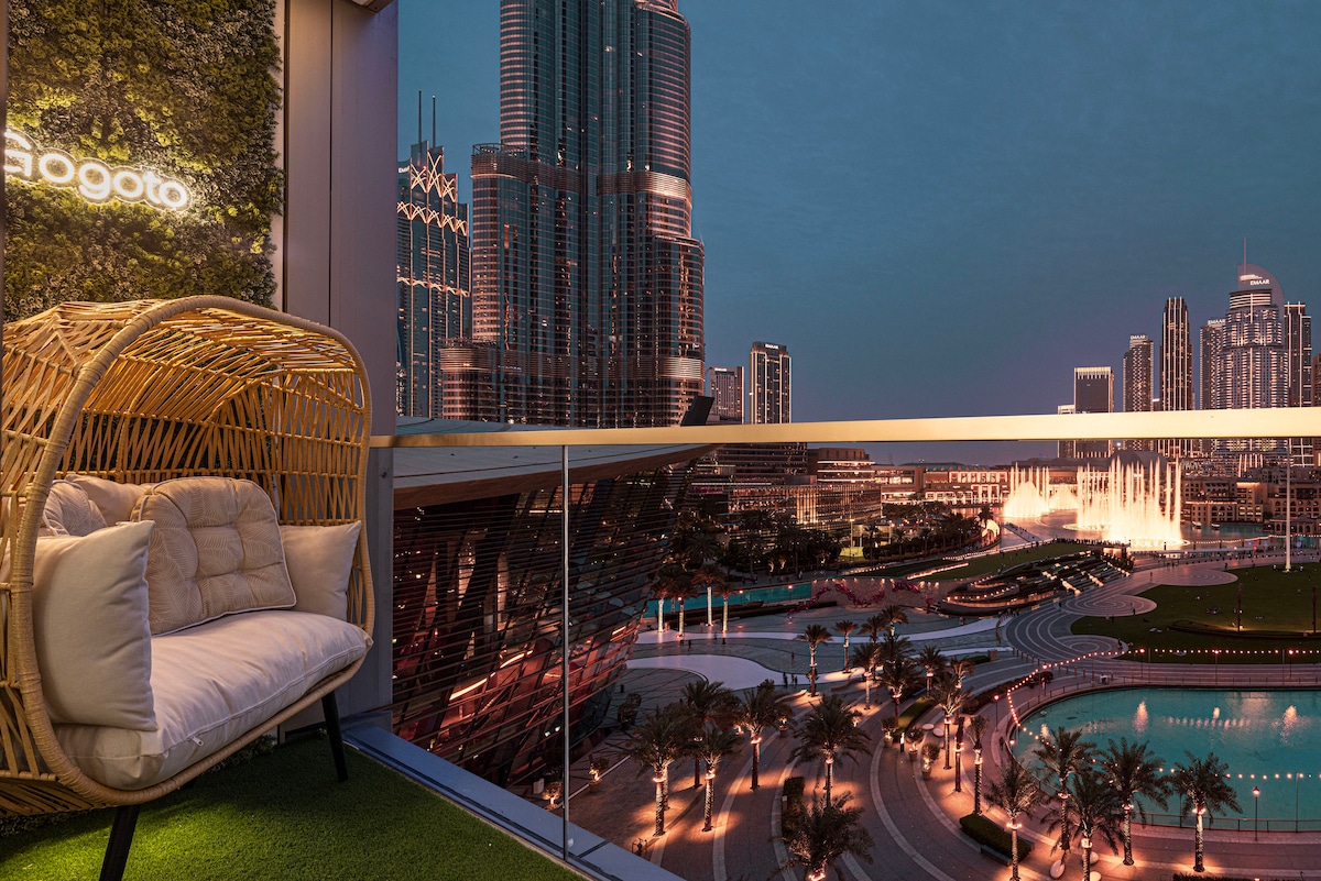 A comfortable seating area is showcased on the balcony, featuring a cozy rattan chair with soft cushions. The view presents a vibrant skyline of Downtown Dubai, highlighted by the illuminated Burj Khalifa and a captivating fountain show in the foreground.