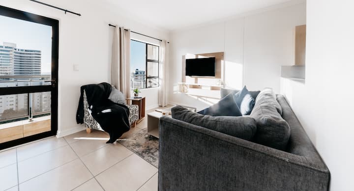 הירגעו בסלון מעוצב עם תוכנית פתוחה הכולל ספה מפנקת בצורת L, כיסא מודגש מודרני וטלוויזיה חכמה 55 אינץ' עם נטפליקס, עיצוב אלגנטי, תאורה רכה ולוח צבעים מרגיע יוצרים את המרחב המושלם להירגע או ליהנות.