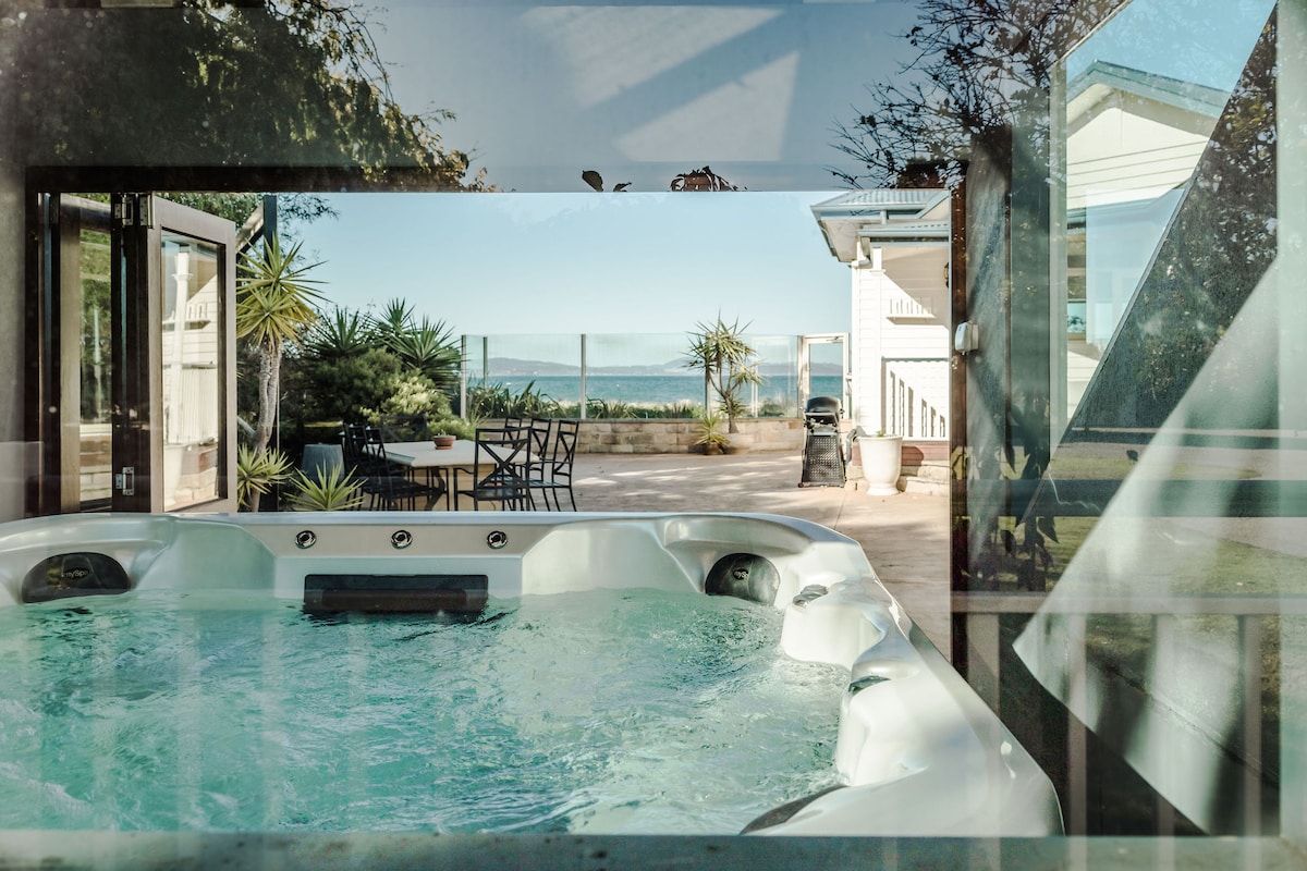 A private hot tub is highlighted in the foreground, surrounded by glass. A view of the outdoor patio reveals seating arrangements and clear vistas of the ocean beyond, framed by tropical plants in the garden.