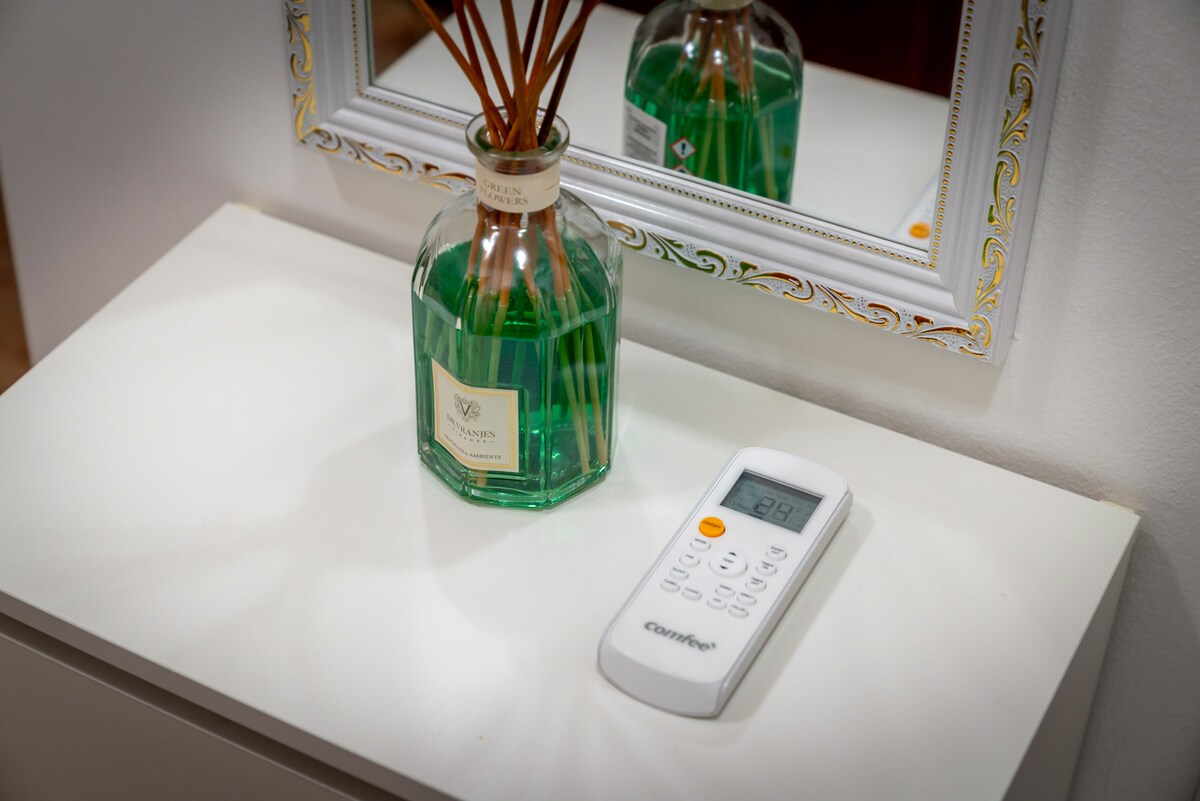 A sleek white remote control is placed on a light-colored surface beside a decorative glass vase containing reed diffusers. A compact mirror with an ornate frame reflects the scene, adding depth to the setting.