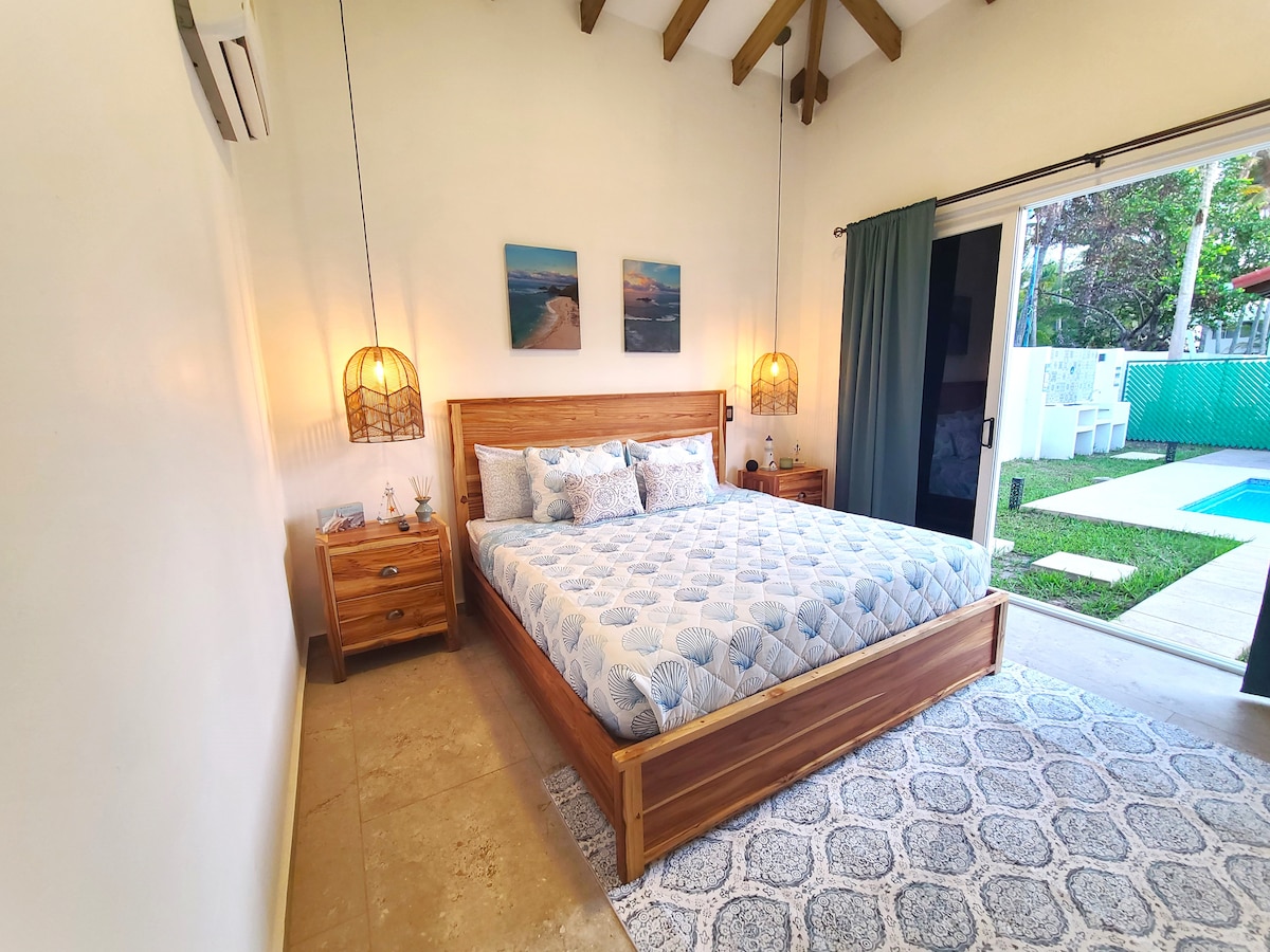 A spacious bedroom is featured, highlighted by a large bed adorned with a light blue patterned blanket. Natural light enters through sliding doors that lead to a garden area with a pool. Wooden nightstands flank the bed, enhancing the warm ambiance.