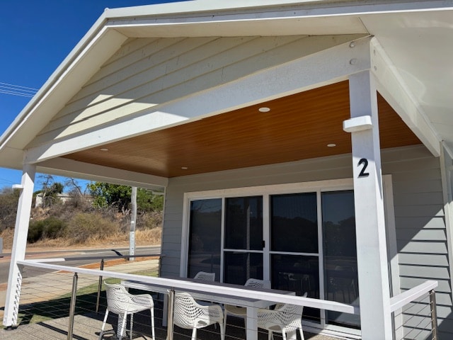 An outdoor patio area features a covered roof with wooden paneling, providing shade. A white table is surrounded by multiple white chairs, creating a comfortable space for dining or relaxation. Large glass doors lead into the interior, enhancing access to outdoor living.