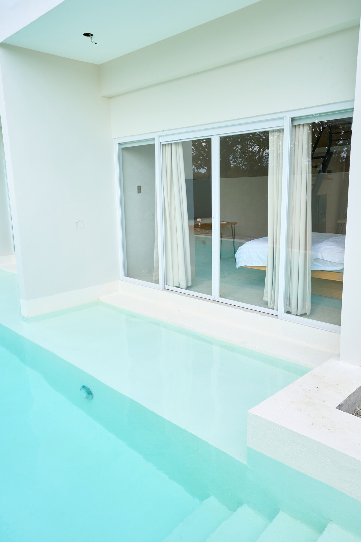 A serene view of a pool area is presented, featuring sliding glass doors that open into a well-lit room. The clear turquoise water reflects the natural surroundings, enhancing the inviting atmosphere. Light-colored curtains gently frame the entrance to the room, creating a seamless indoor-outdoor connection.