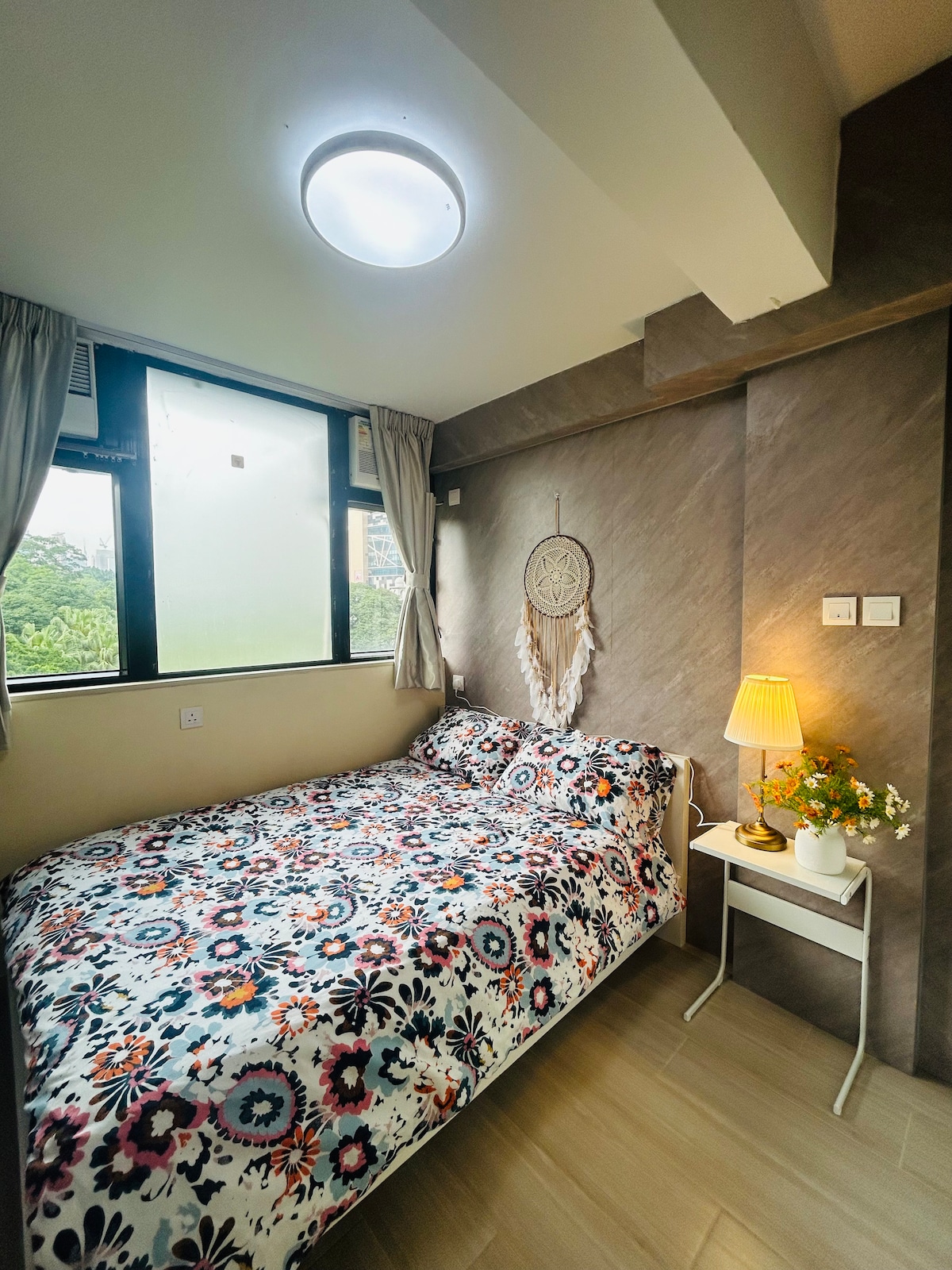A well-lit bedroom features a spacious bed covered with a floral patterned bedspread. Natural light enters through large windows with light curtains. A bedside table holds a lamp and a small vase with flowers, adding a touch of color to the neutral walls.