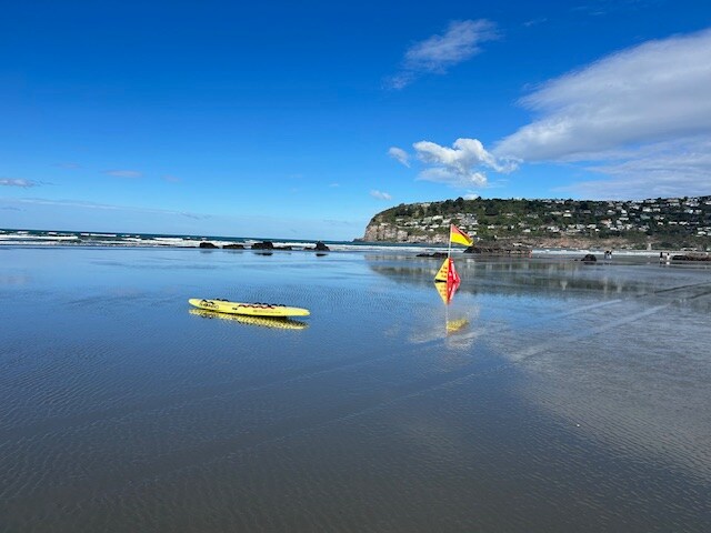 A wide sandy beach extends towards the ocean, reflecting the clear blue sky. A yellow kayak rests on the wet sand, and colorful flags indicate safety zones. A coastal hill with greenery rises in the background, enhancing the tranquil seaside environment.