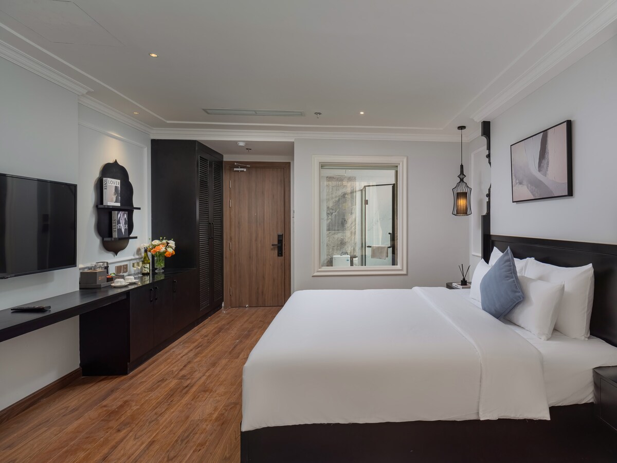 A well-appointed guest room features a spacious bed with crisp white linens and a decorative blue pillow. A large window provides natural light, while a dark wooden desk and television enhance the modern decor. Subtle artwork and a warm wood floor complete the inviting atmosphere.