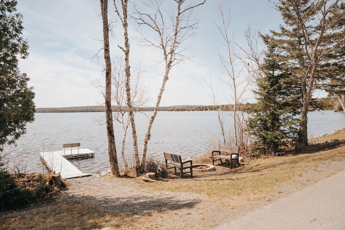 The scene features a serene lakeside area, with a wooden dock extending into the water. Benches are positioned nearby, surrounded by trees and natural greenery. A fire pit can be seen, providing an inviting space for gatherings. The calm water reflects the clear sky.