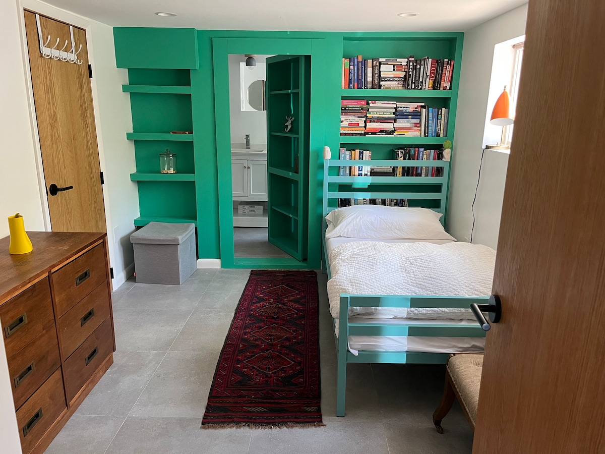 A cozy sleeping area is defined by a twin bed framed in teal. A colorful bookshelf is positioned against the wall, filled with various books. A dresser stands nearby, and a decorative rug adds warmth to the tiled floor. A doorway leads to an en suite bathroom.