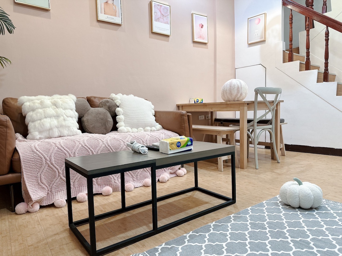 A cozy living area features a brown sofa adorned with soft, textured pillows and a light pink throw. A small coffee table sits in front, complemented by a dining table and chairs. The pastel walls are decorated with framed art, and a staircase is visible in the background.