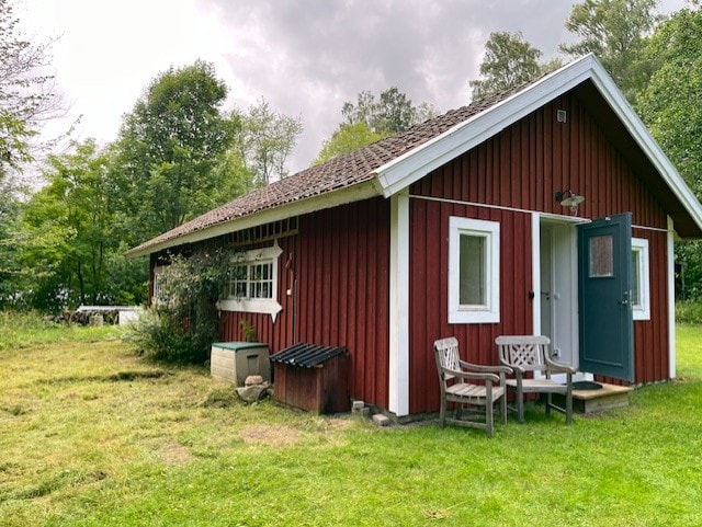 A cozy red cottage is nestled within a grassy area, surrounded by trees. Two chairs are positioned on the porch, offering a view of the lush greenery. A small storage unit is visible beside the entrance, enhancing the rustic charm of the setting.