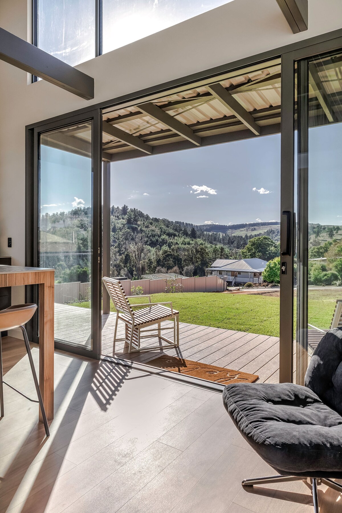 The image captures a bright, open space with large glass doors leading to a deck area. A single chair is positioned on the deck, overlooking a grassy yard and distant hills under a clear sky, inviting outdoor relaxation and scenic views.