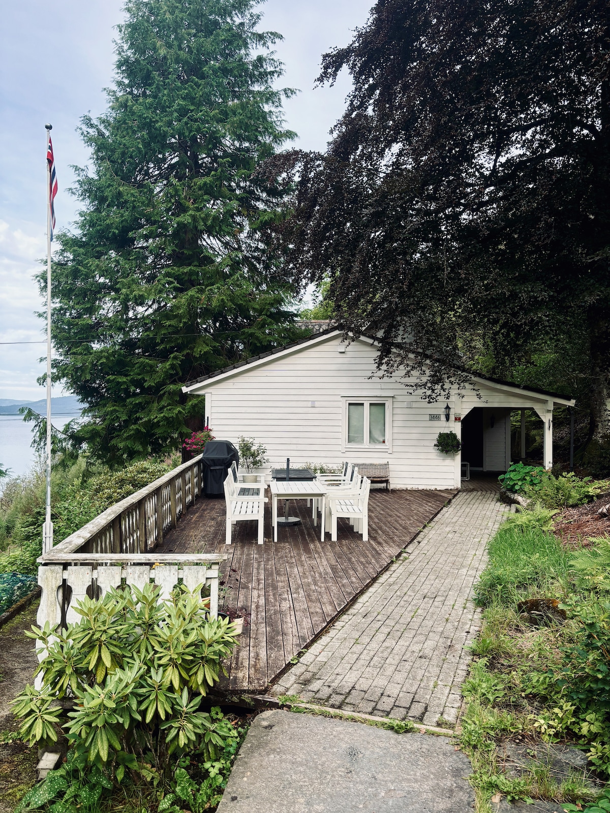 The exterior of a white house is shown, surrounded by lush greenery and tall trees. A wooden deck features a dining table and chairs, offering a tranquil view of the water. A pathway leads to the entrance, enhancing accessibility to the home.