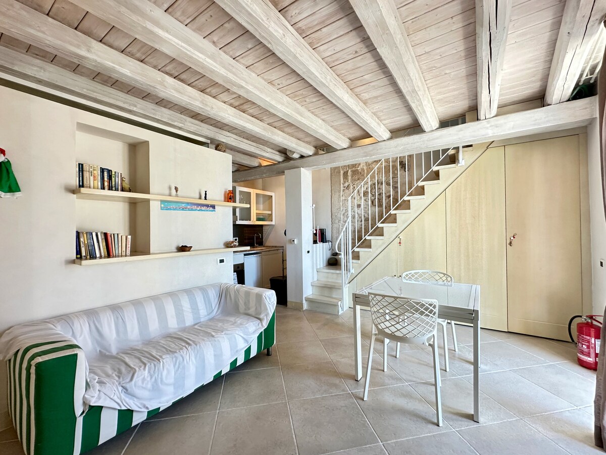 A spacious living area features a white sofa with green stripes, an open kitchen, and a dining table surrounded by white chairs. A staircase leads to an upper level. Wooden beams complement the neutral-toned walls, while a bookshelf displays various books.
