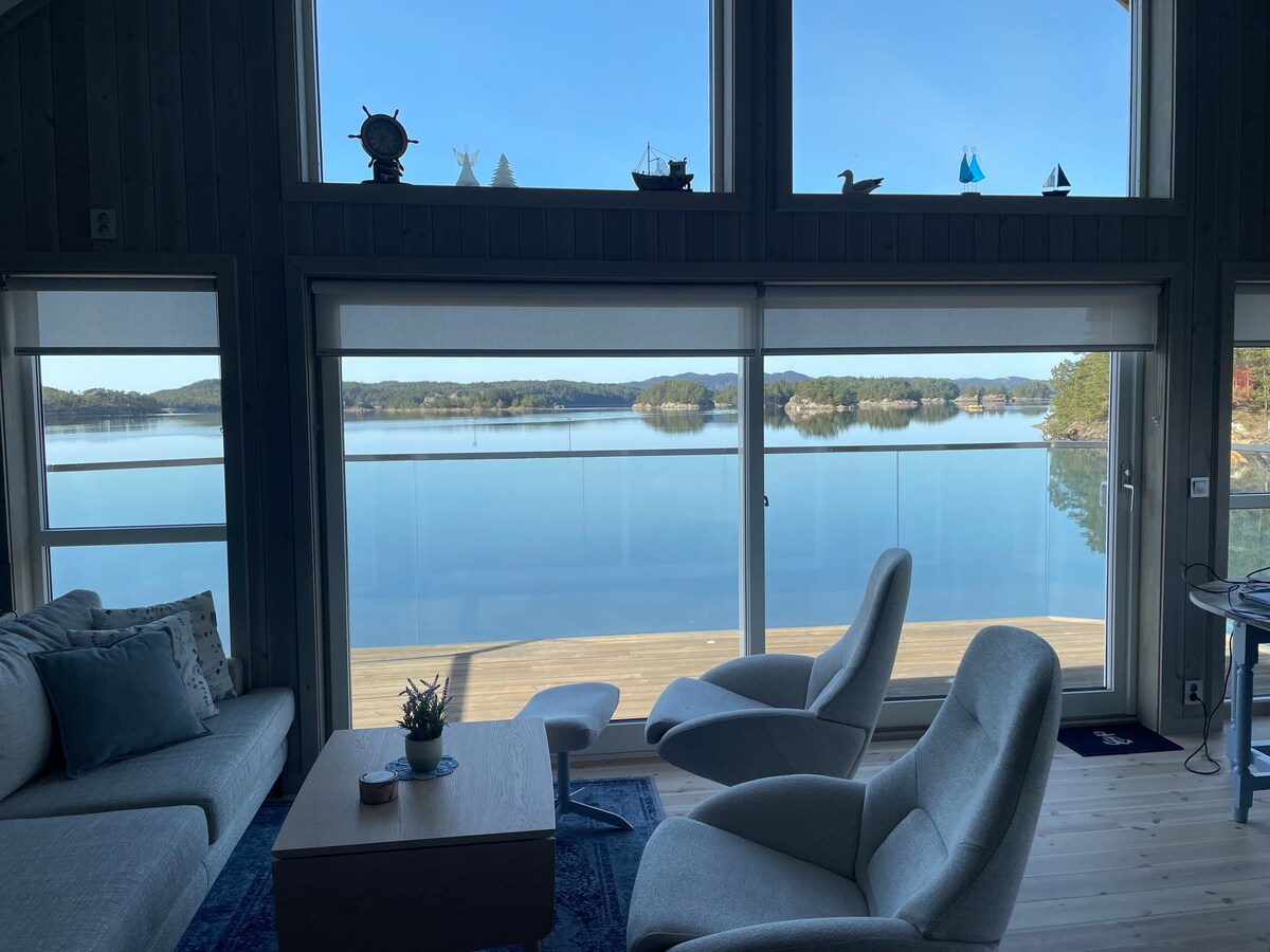 A spacious living area features large windows offering unobstructed views of the calm waters and surrounding landscape. Comfortable seating is provided by two light gray armchairs and a soft couch, complemented by a coffee table. Natural light filters in, enhancing the relaxing ambiance.