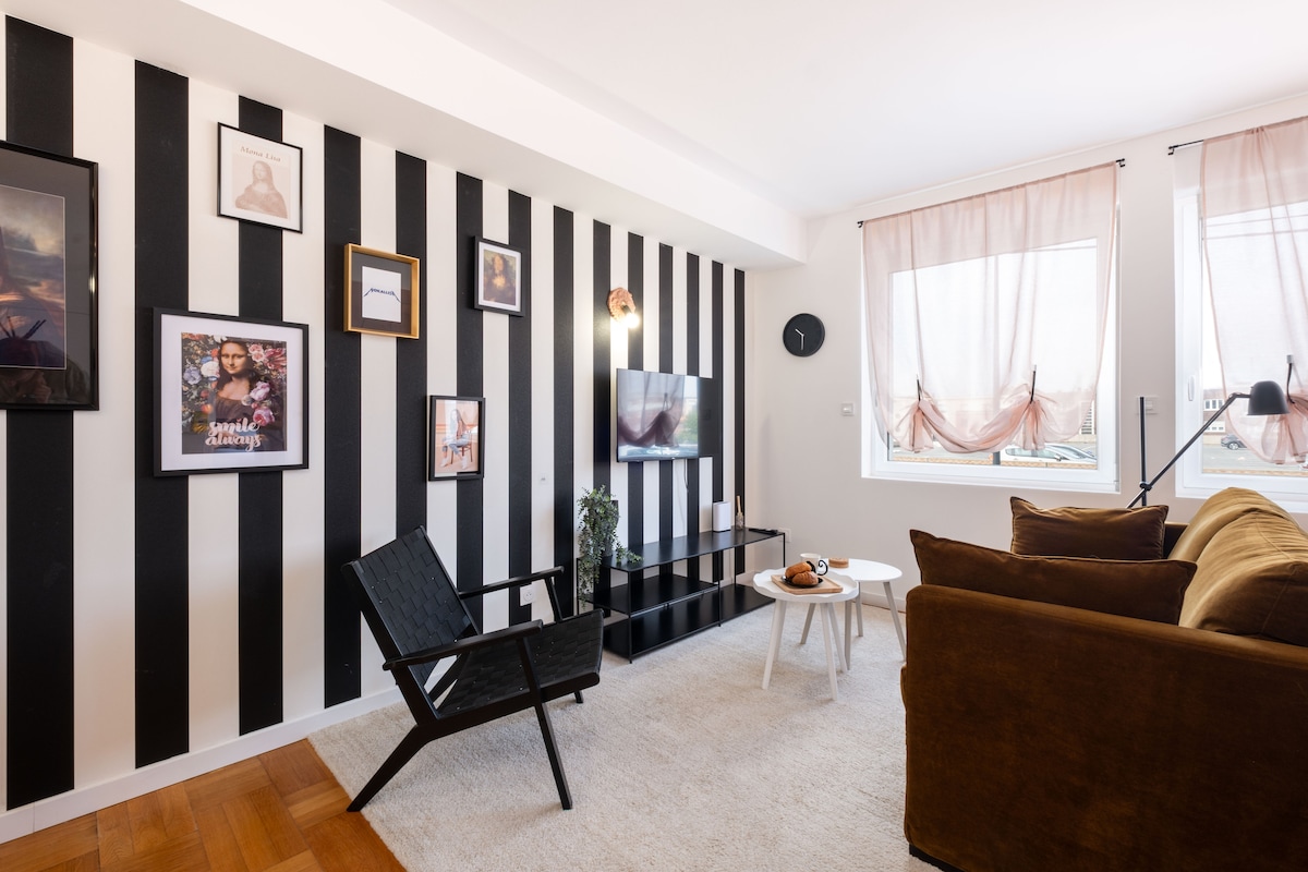 A stylish living space features a black and white striped wall, complemented by two large windows adorned with light fabric curtains. A relaxed seating area includes a brown sofa and a sliding black media unit, with framed artwork and a small coffee table showcasing decorative items.