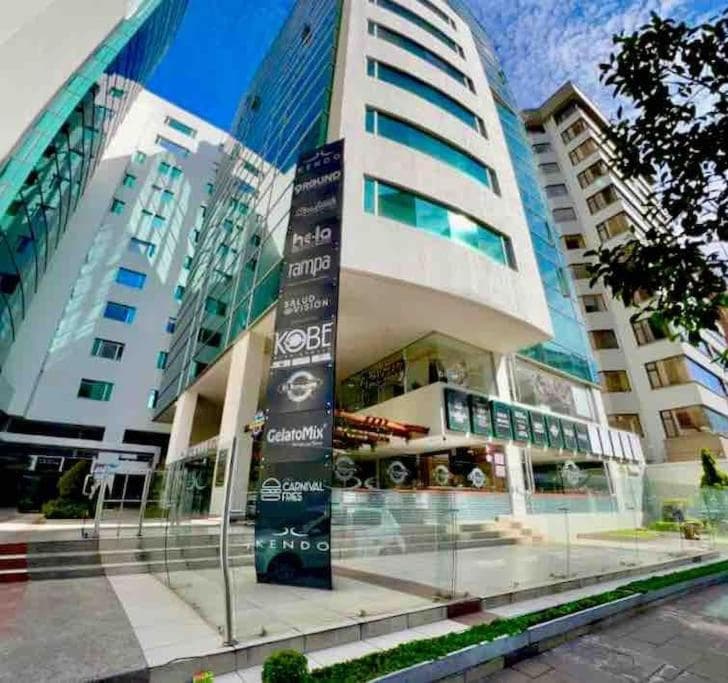 The exterior of Kendo Plaza is characterized by its modern architecture, featuring large glass windows and a sleek design. A welcoming entrance is framed by greenery and outdoor seating areas, with clear signage indicating various businesses located within the building.