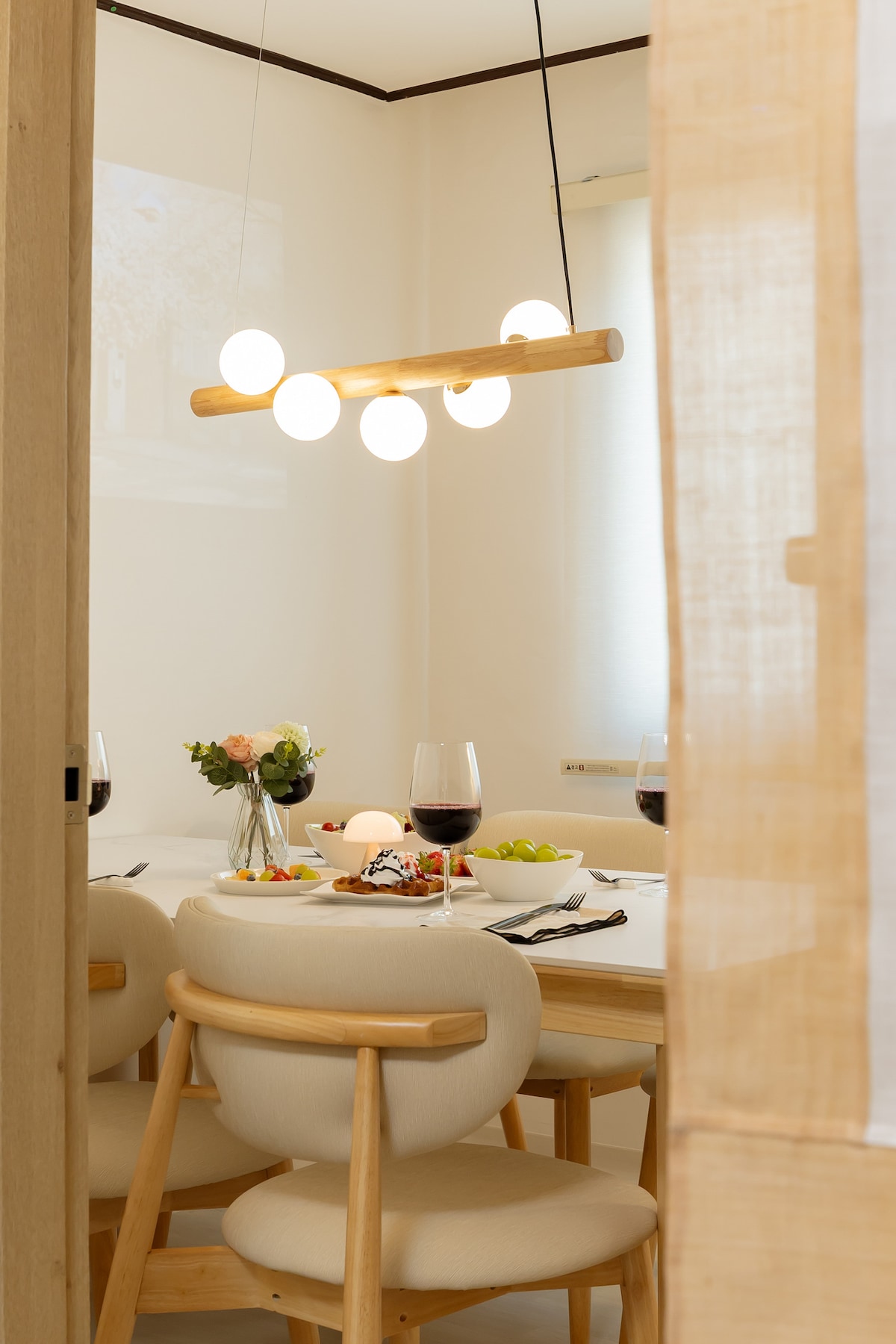 A dining area is presented with a light wooden table set for a meal. Six upholstered chairs surround the table, featuring simple decor and soft lighting from a modern overhead fixture. A vase with flowers and dishes are neatly arranged on the table, providing an inviting atmosphere.