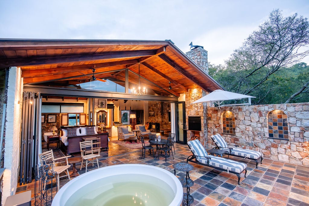 An open outdoor area showcases a spacious hot tub surrounded by comfortable loungers and dining chairs. The roof's wooden beams enhance the rustic charm of the stone-walled space, which is illuminated by hanging lights. A cozy lounge area is visible under the shelter.